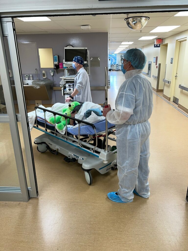 Parent (white jumpsuit) helping nurses guide patient to the operating room. Some parents escort right to the operating suite or while others leave at the threshold of the operating room entrance.